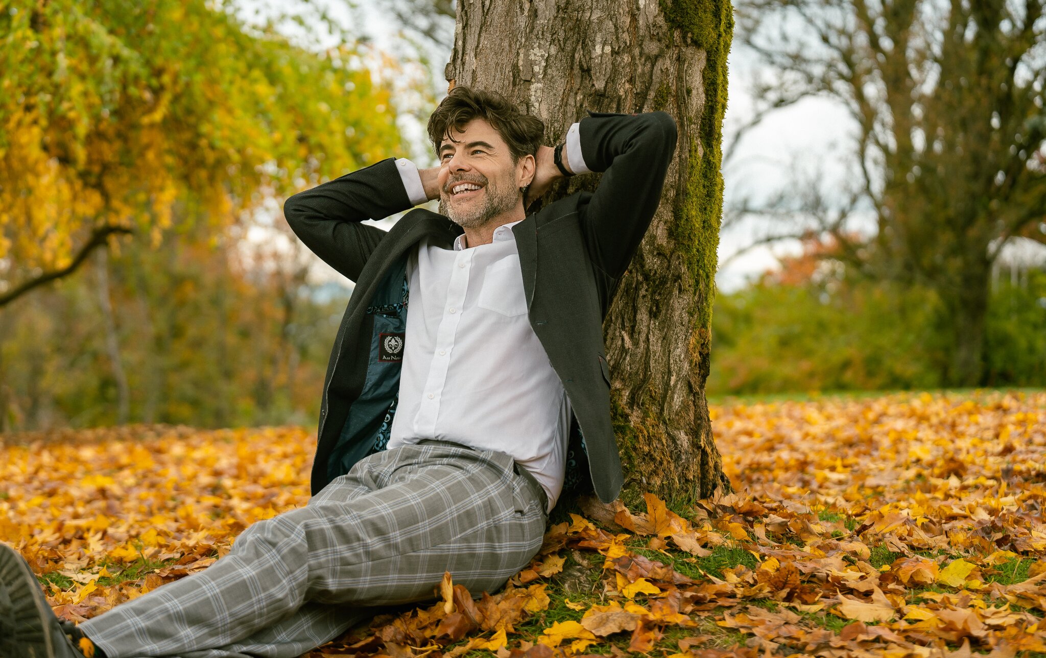 Man in a suit leans against a tree, smiling with hands behind head, surrounded by colorful autumn leaves. The scene is peaceful and joyful.