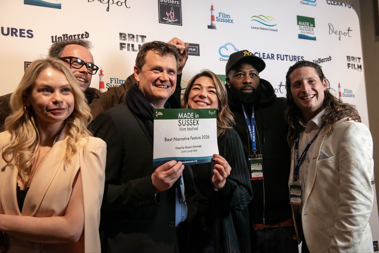 Group of six people smiling and holding an award plaque at a film festival event. The backdrop features various sponsor logos.