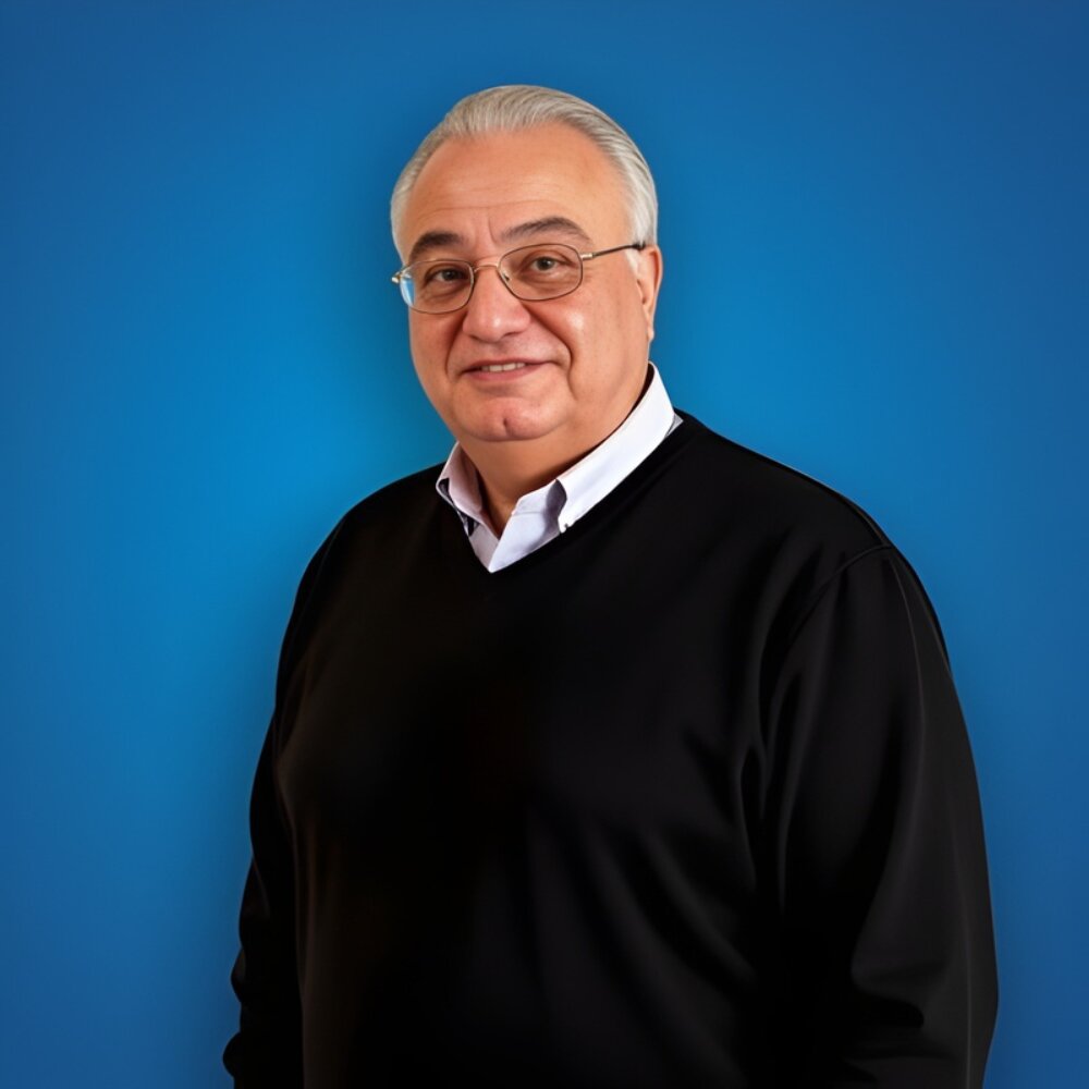 A smiling older adult with glasses stands against a solid blue background. He's wearing a black sweater over a white shirt, exuding a friendly and approachable demeanor.