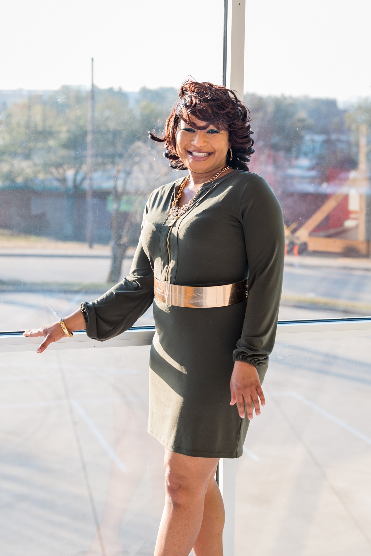 Close-up of smiling woman in green dress with gold belt standing by window.