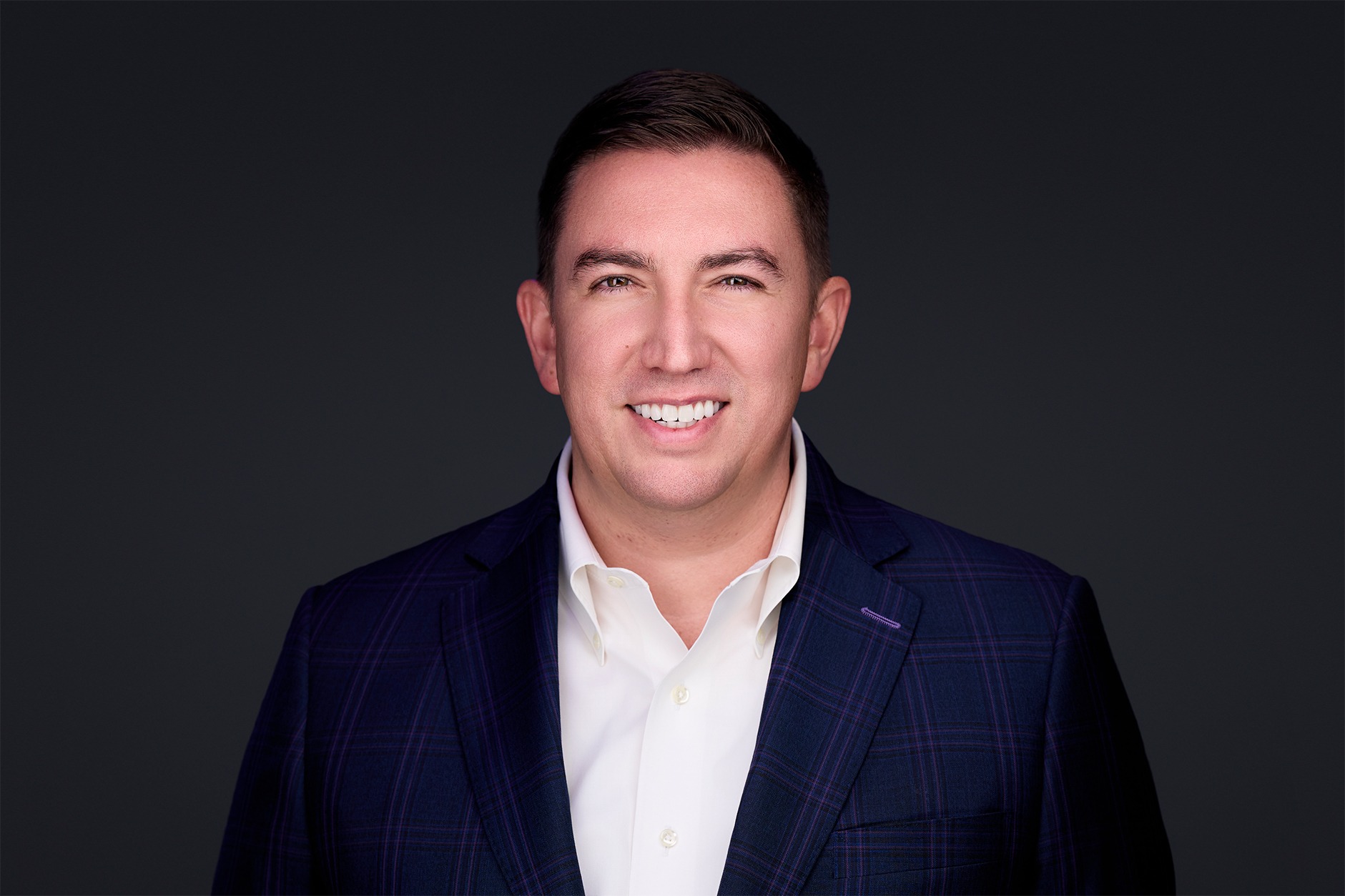 Smiling man in a dark plaid suit jacket and white shirt against a gray background. The expression is confident and friendly.
