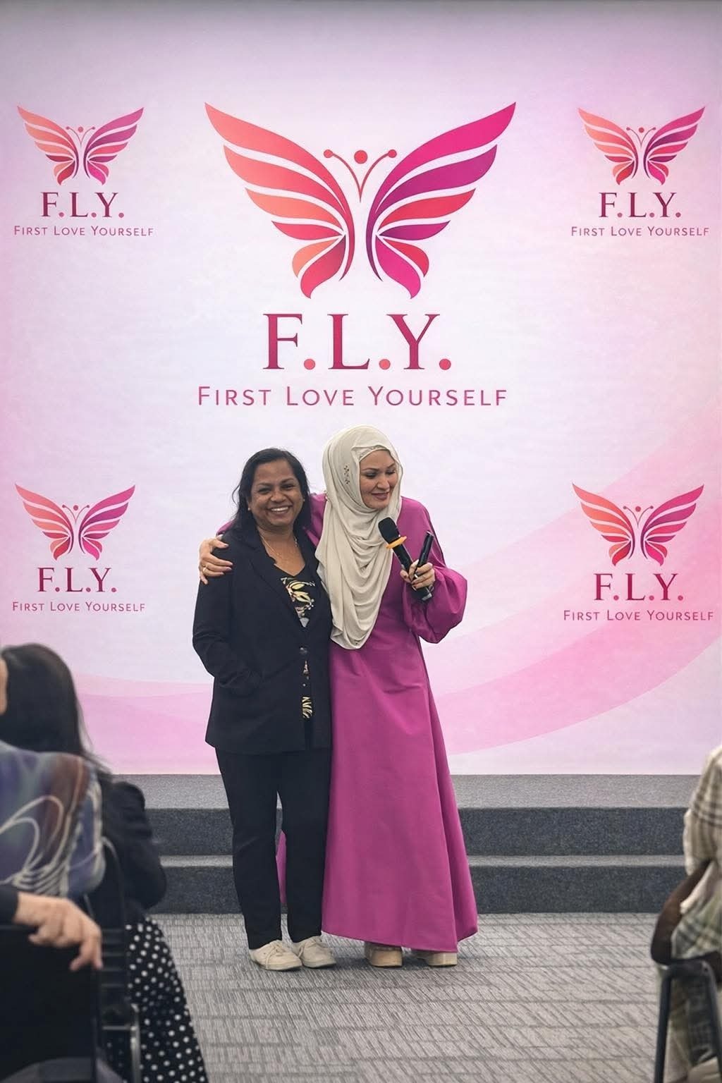 Two women smiling on stage at FLY event backdrop, one in pink dress holding microphone.