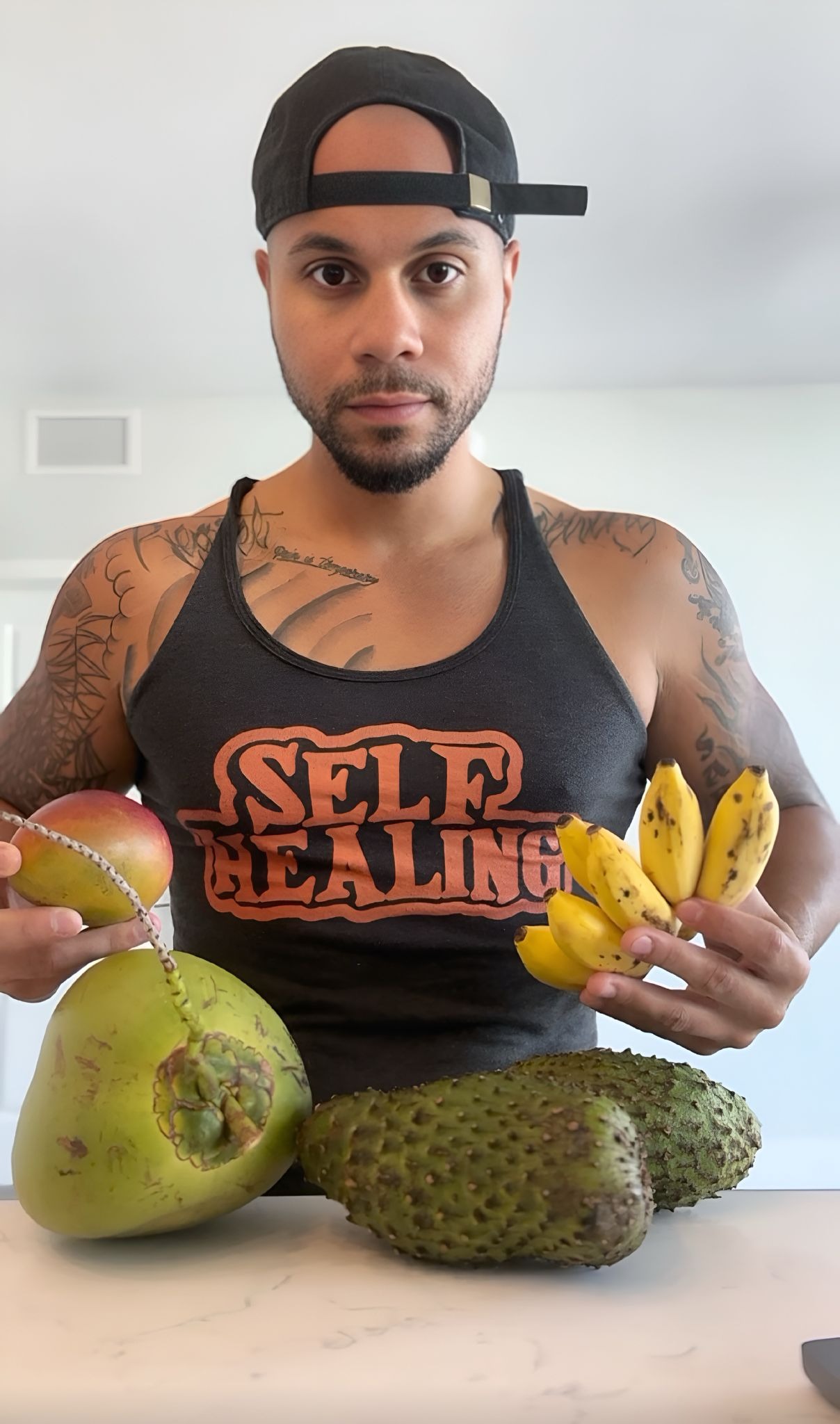 A person in a black tank top and cap holds tropical fruits—mango, mini bananas, coconut, and soursop—conveying a fresh and healthy vibe.