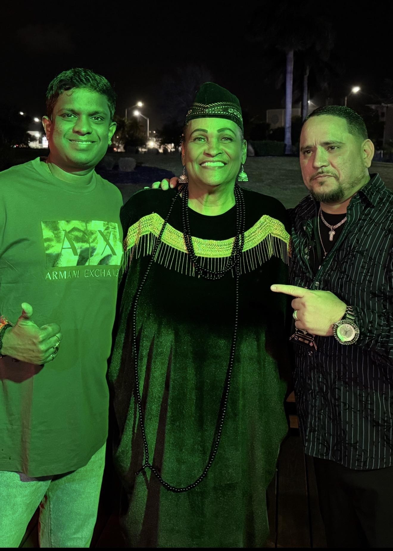 Three people posing at night under green lighting, woman in traditional attire, men beside her smiling and pointing.