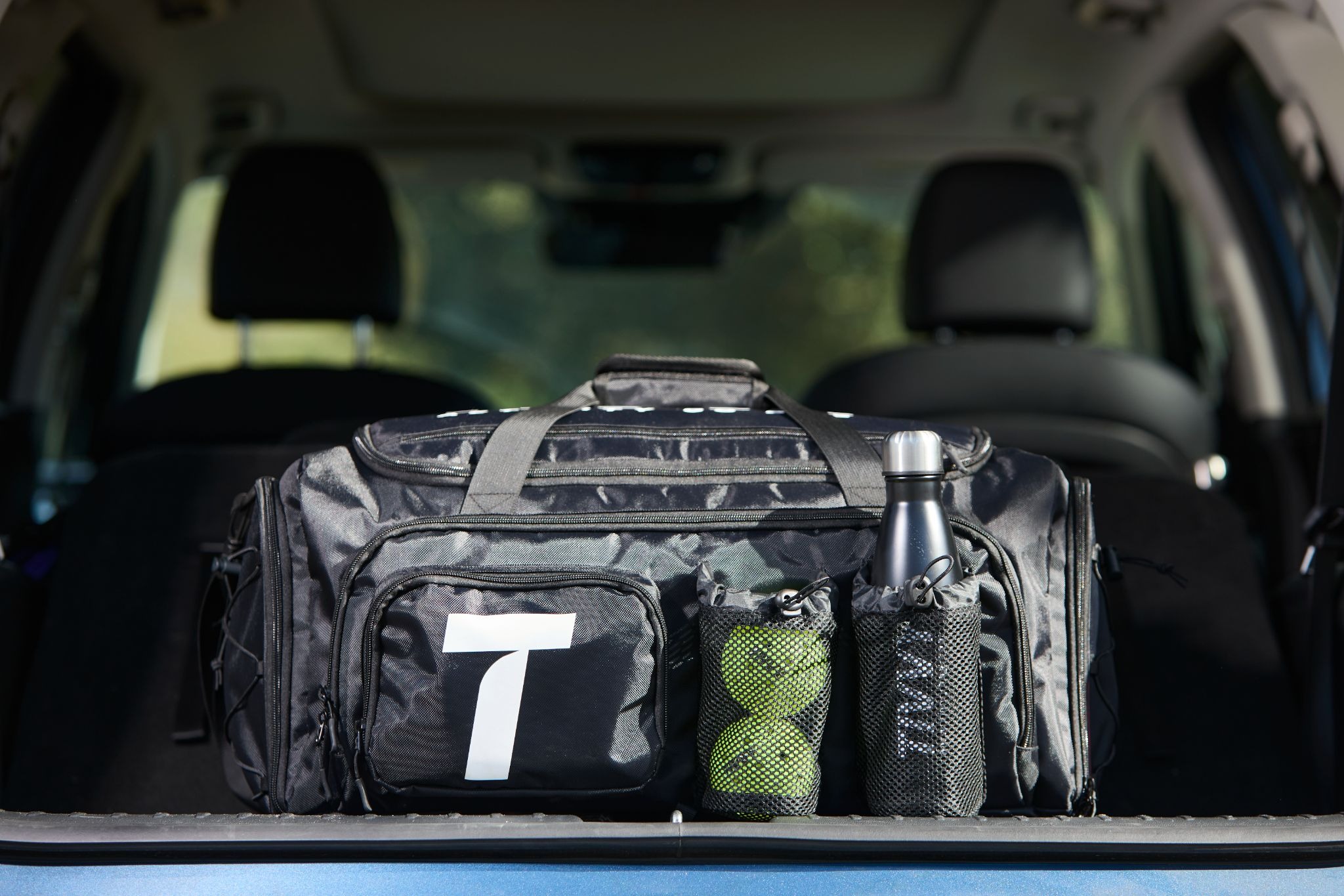 Black sports bag in a car trunk, featuring mesh pockets with tennis balls and a water bottle. The scene suggests readiness for outdoor activities.
