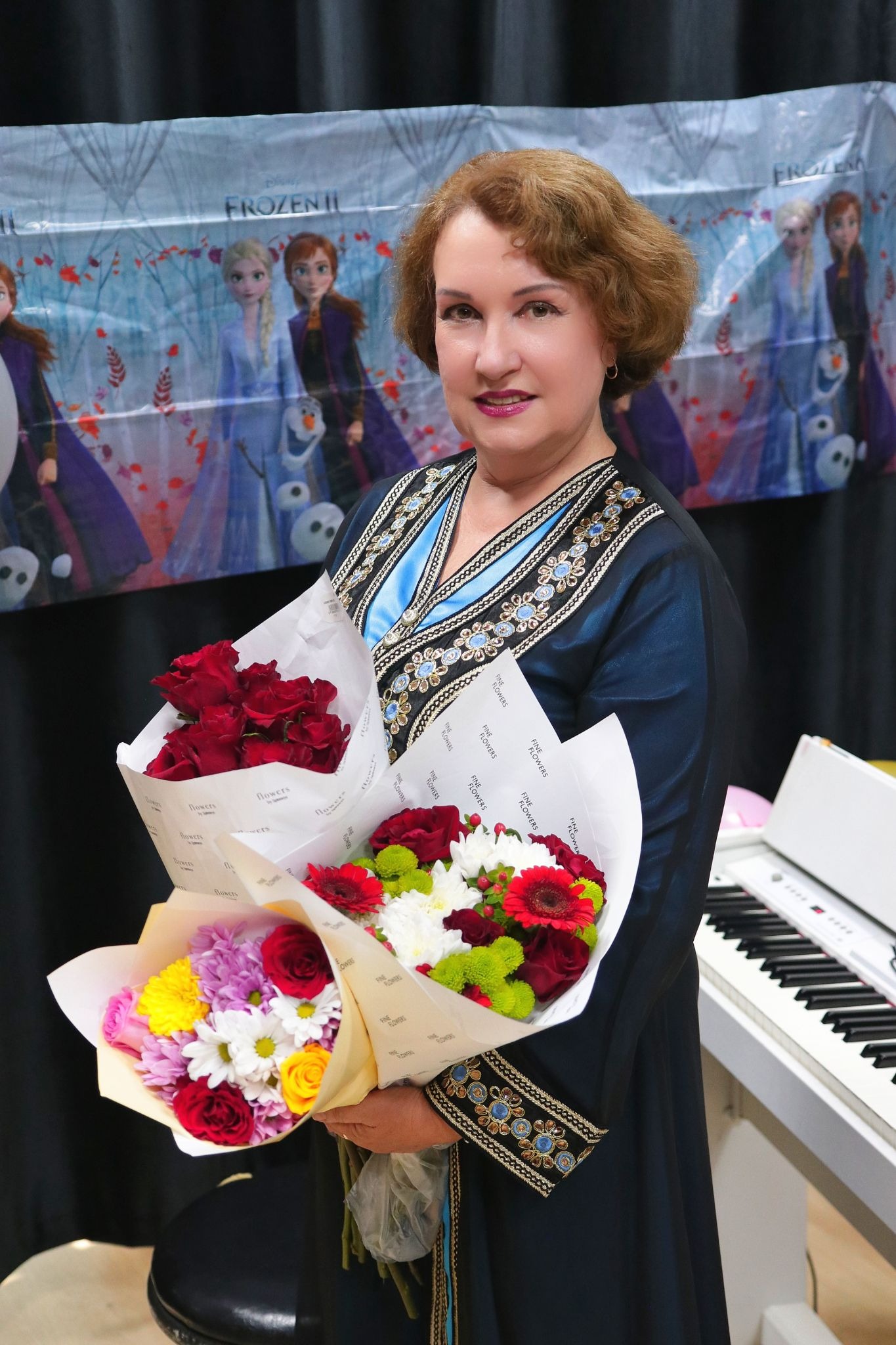 A smiling woman in an ornate blue dress holds two bouquets of colorful flowers. A "Frozen II" backdrop and a piano keyboard are visible behind her.