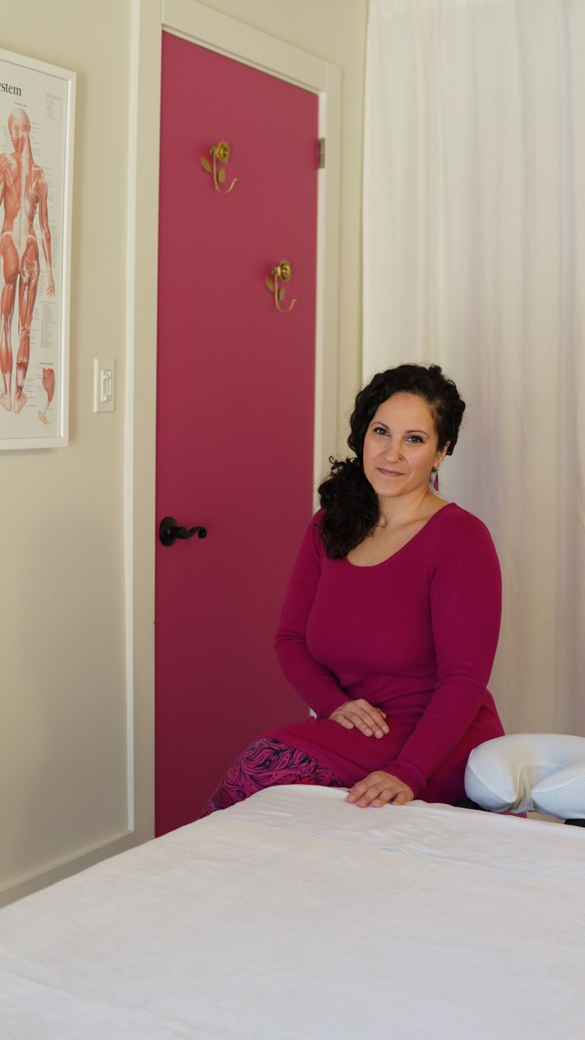 Woman in a magenta top sits in a massage room, with a chart of the muscular system on the wall and a magenta door. Calm and welcoming atmosphere.