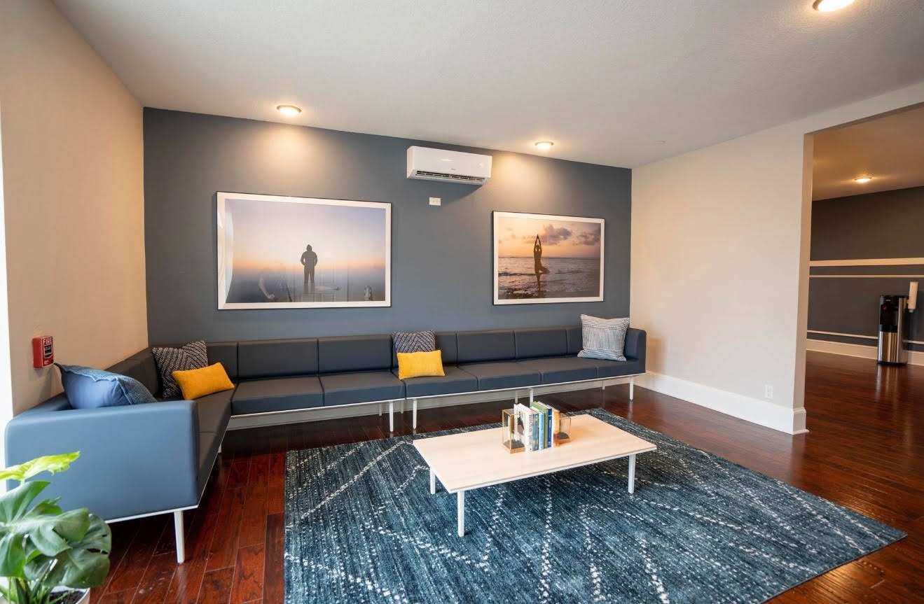 Modern lounge with blue walls, a long gray couch, yellow and gray pillows, coastal photos, a wood coffee table, and a textured blue rug.