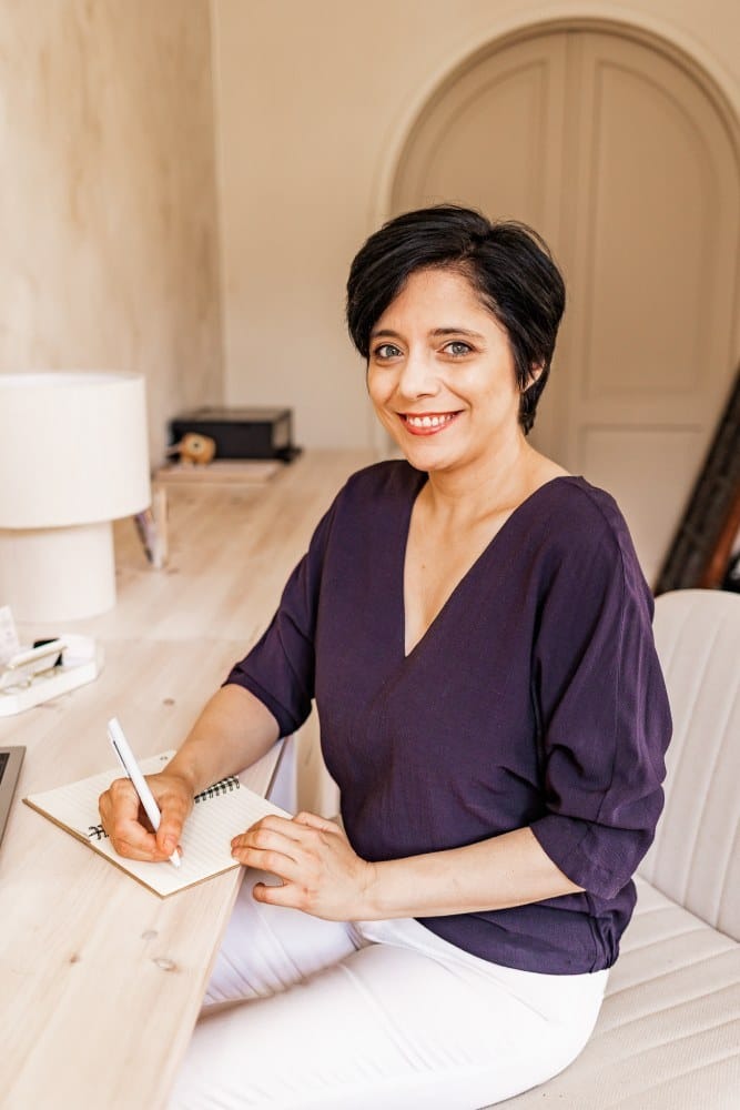 Woman sitting at desk, writing in notebook, smiling in bright minimalist workspace.