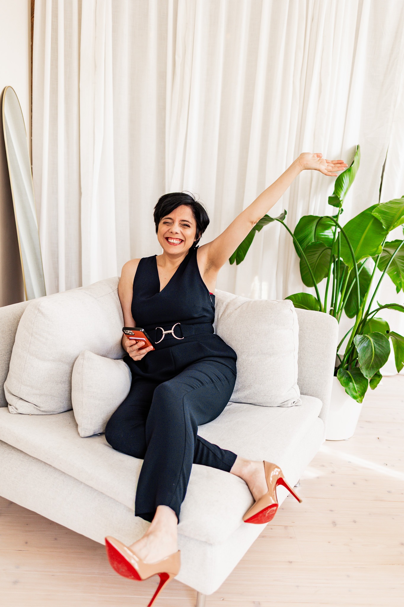 Woman in black outfit relaxing on sofa, holding phone, raising arm joyfully near indoor plant.