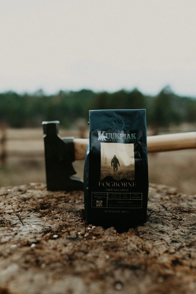 A package of "Fogborne" coffee stands on a chopped tree stump beside an axe in a rustic, misty forest setting, evoking a rugged, adventurous feel.