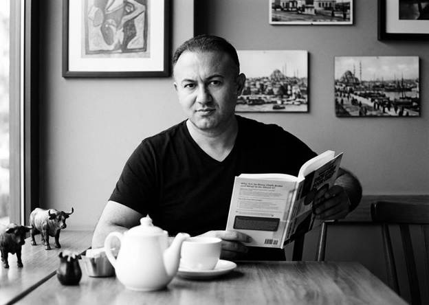 Yusuf Yaran reading Rich Chef Poor Chef at café table with teapot and framed artwork behind him.
