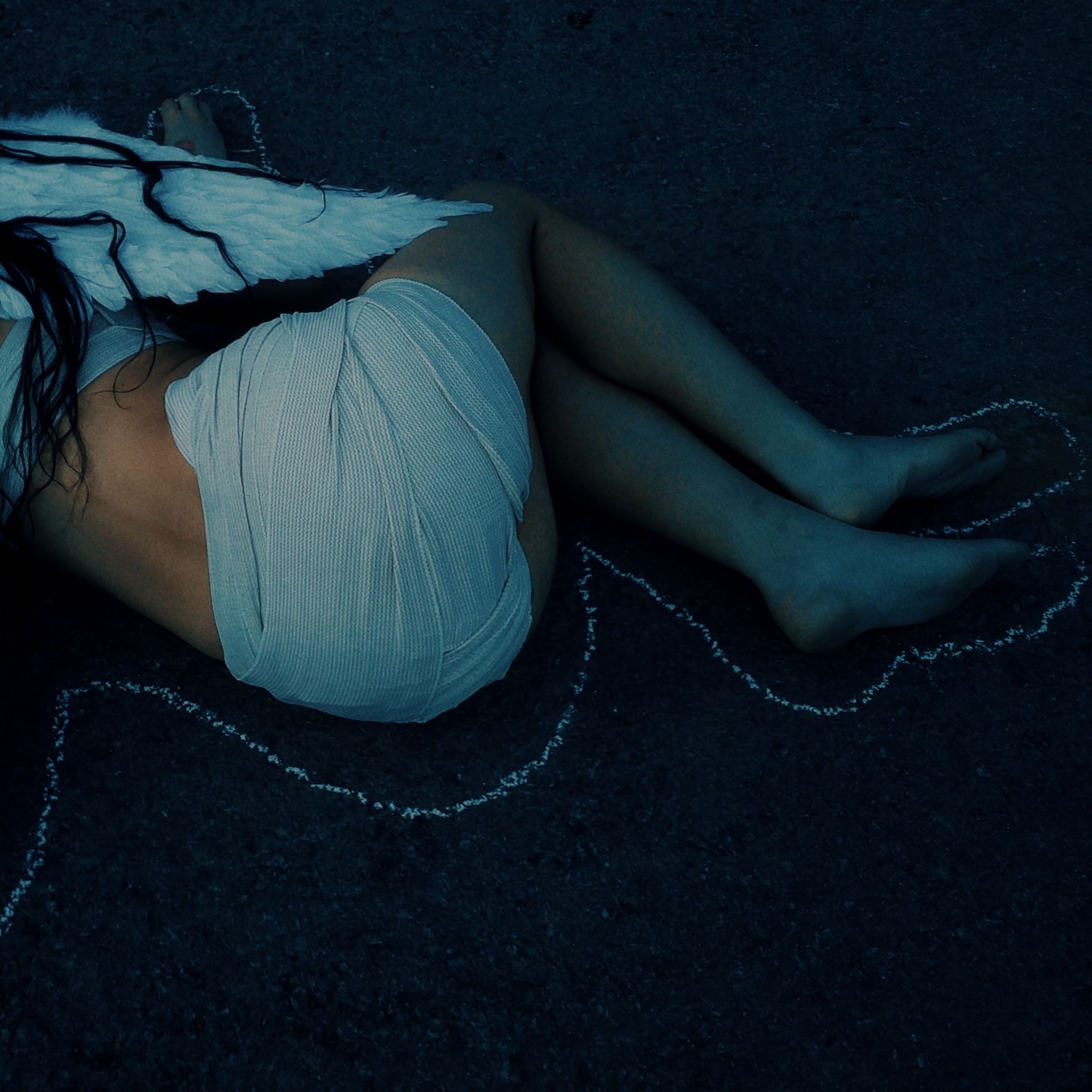 Person with angel wings curled on dark pavement inside chalk outline at night.