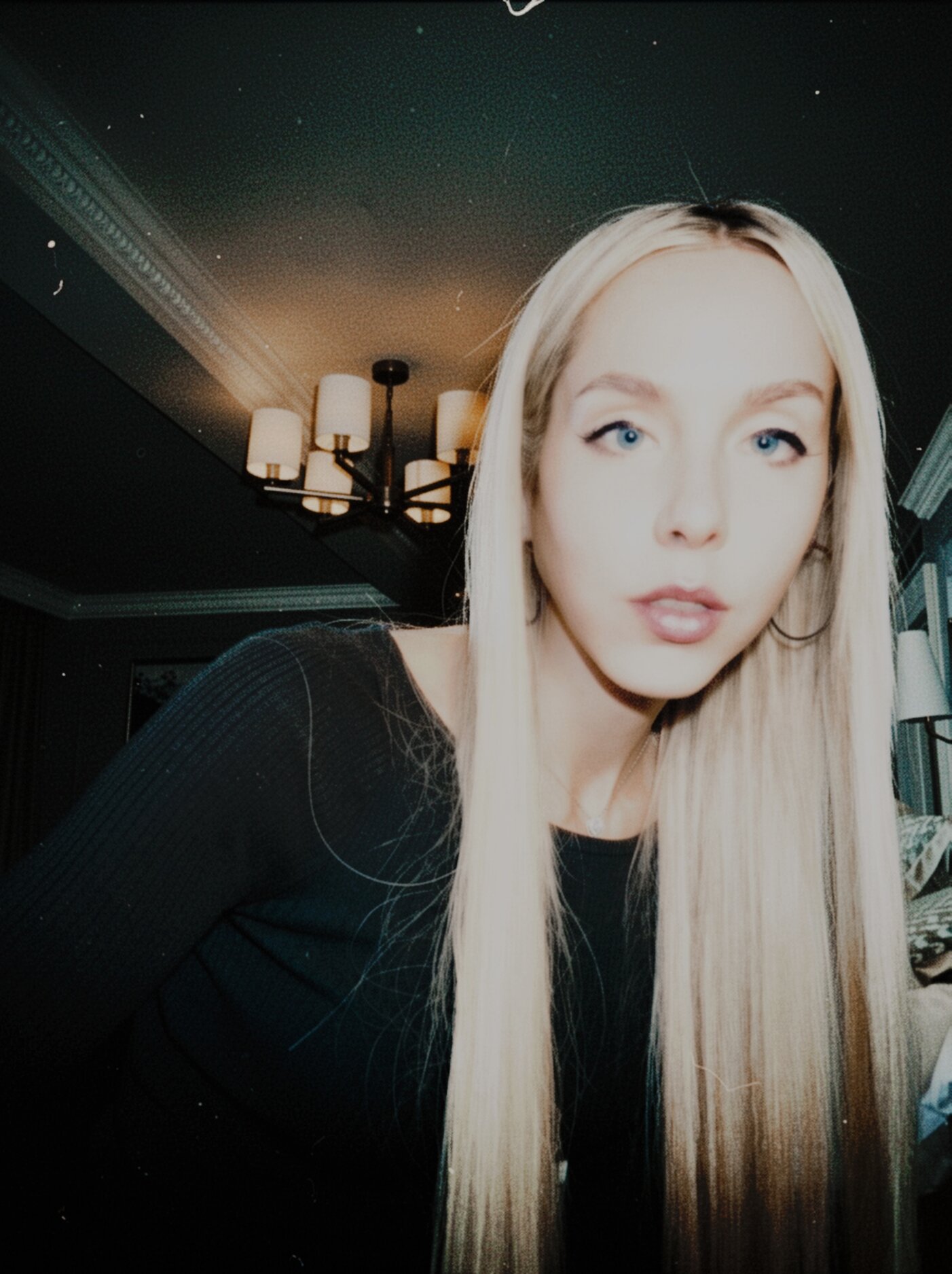 Blonde woman in black top posing indoors beneath chandelier lighting with moody, grainy film aesthetic.