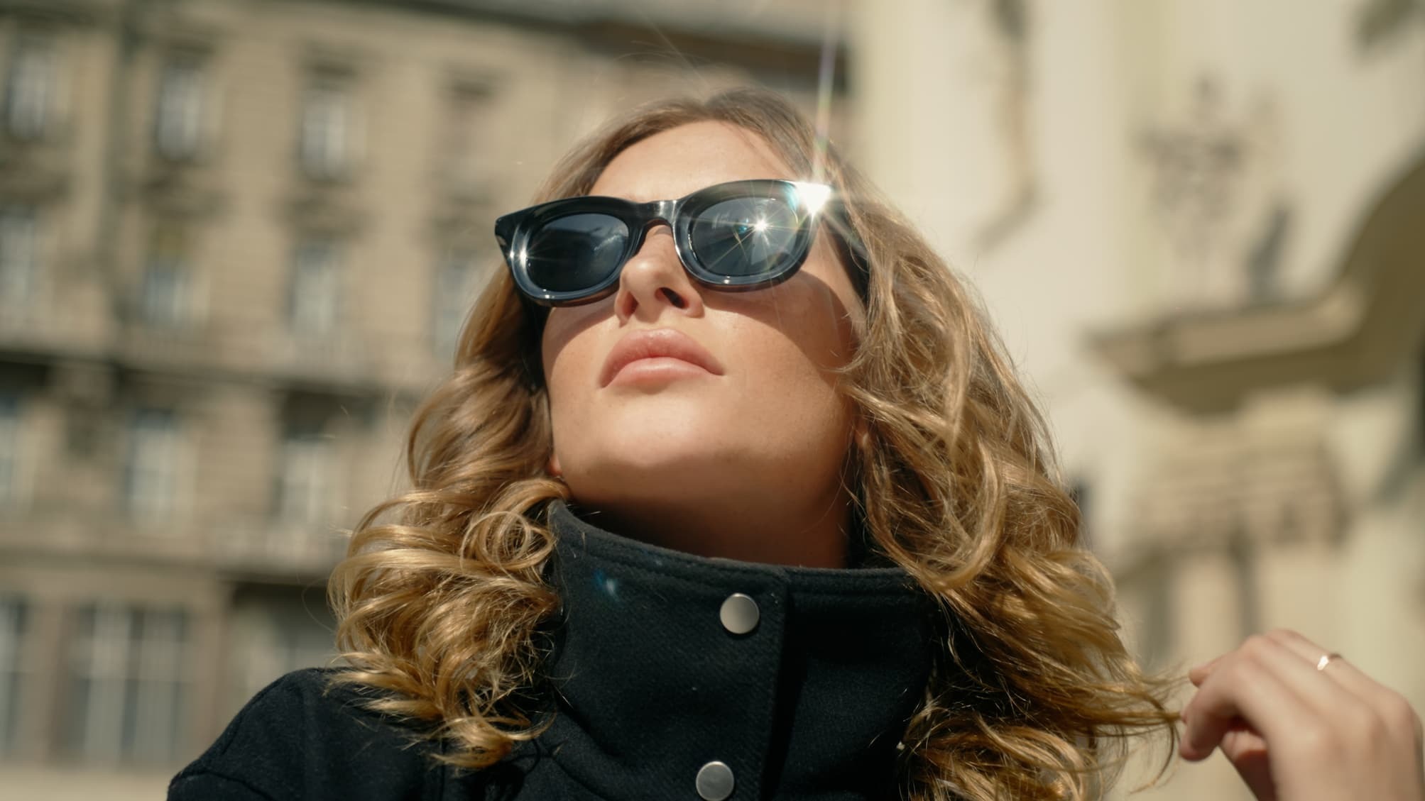 Woman with curly hair wearing sunglasses and a black coat looks upward confidently. Sunlight reflects off her glasses. Urban background in soft focus.