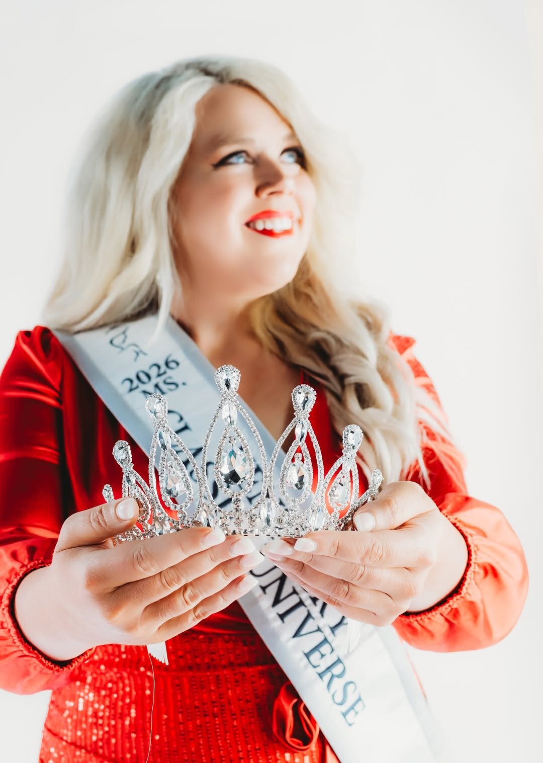 A smiling woman with blonde hair holds a sparkling tiara in her hands. She wears a red dress and a sash, conveying pride and celebration.