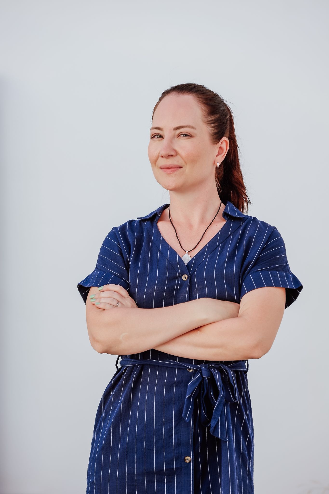 Woman in blue striped dress standing confidently with arms crossed against white background.