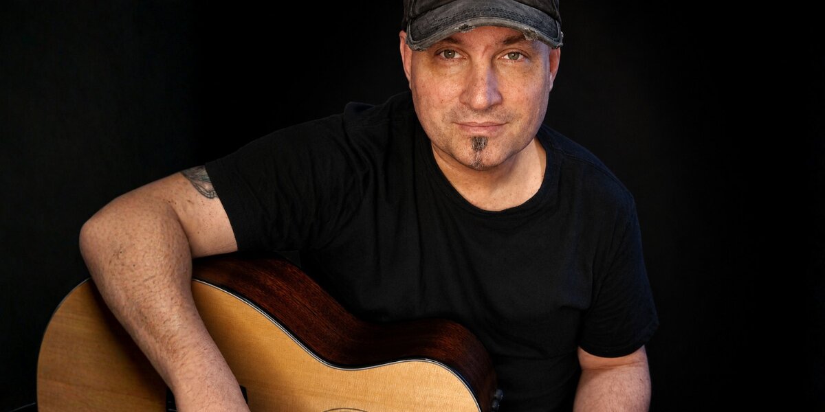Gene Dozois holding an acoustic guitar against a dark background during a promotional portrait.