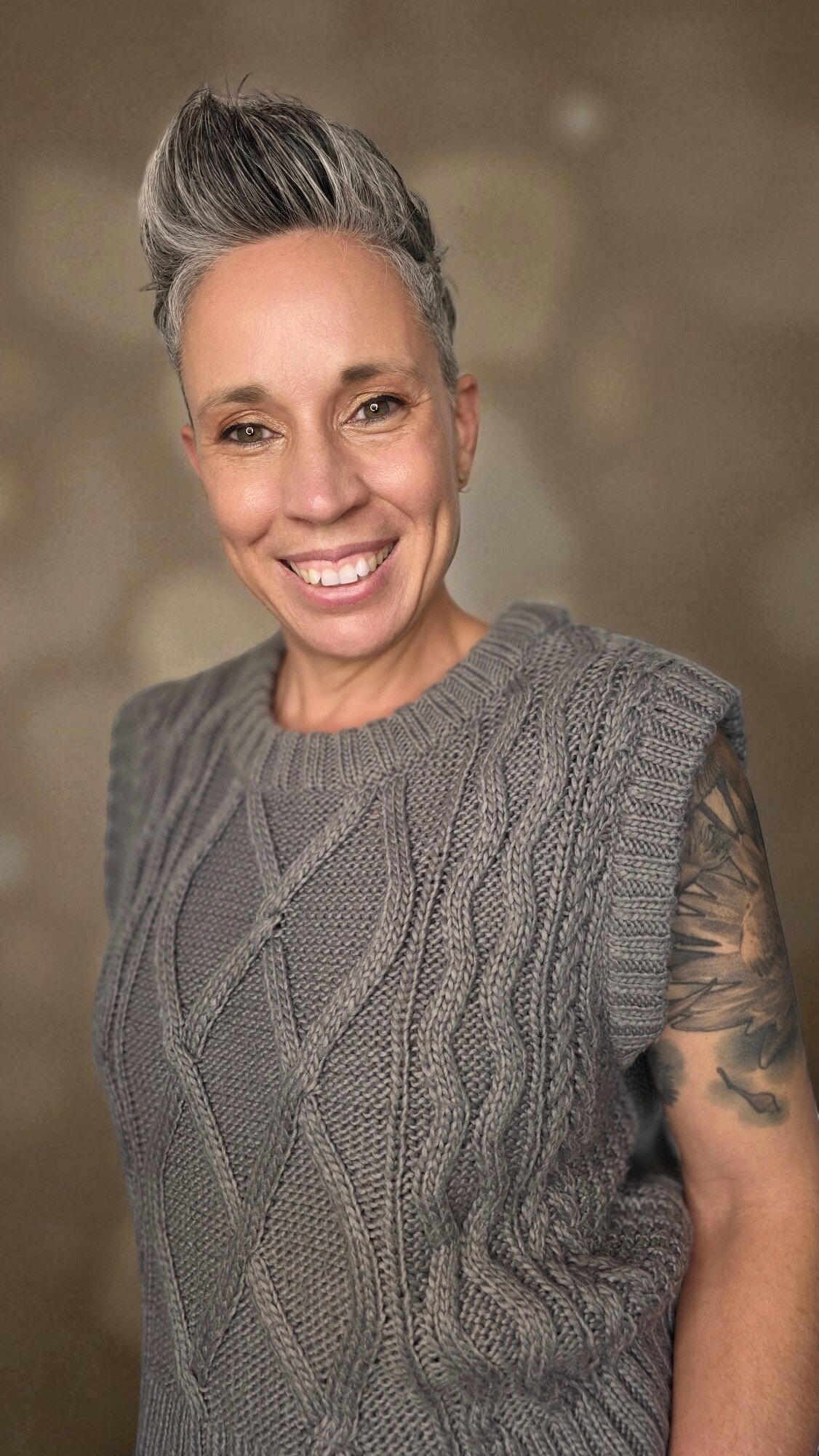 Smiling woman with short gray hairstyle wearing textured gray knit top against soft neutral background.