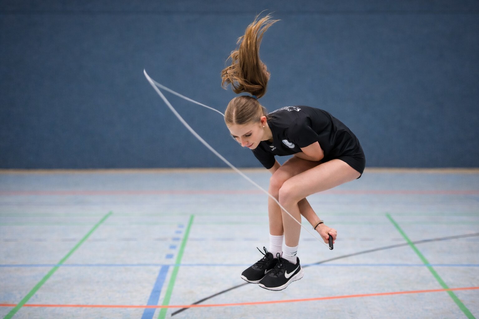 A young woman skillfully performs a jump rope trick in a sports hall, her hair and the rope in motion. The scene conveys energy and focus.