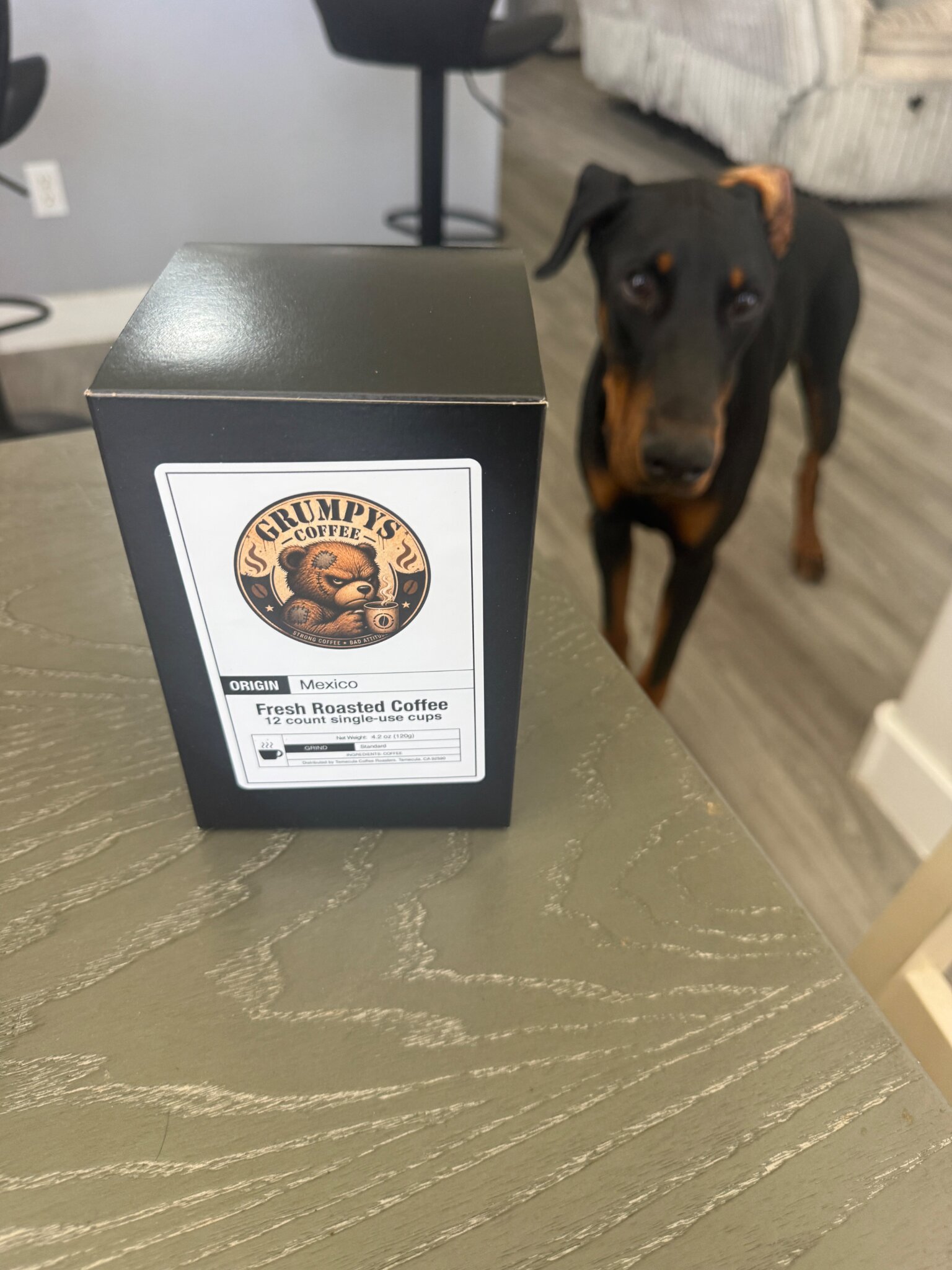 Black Grumpy’s Coffee box on table with Doberman standing nearby in modern home interior.