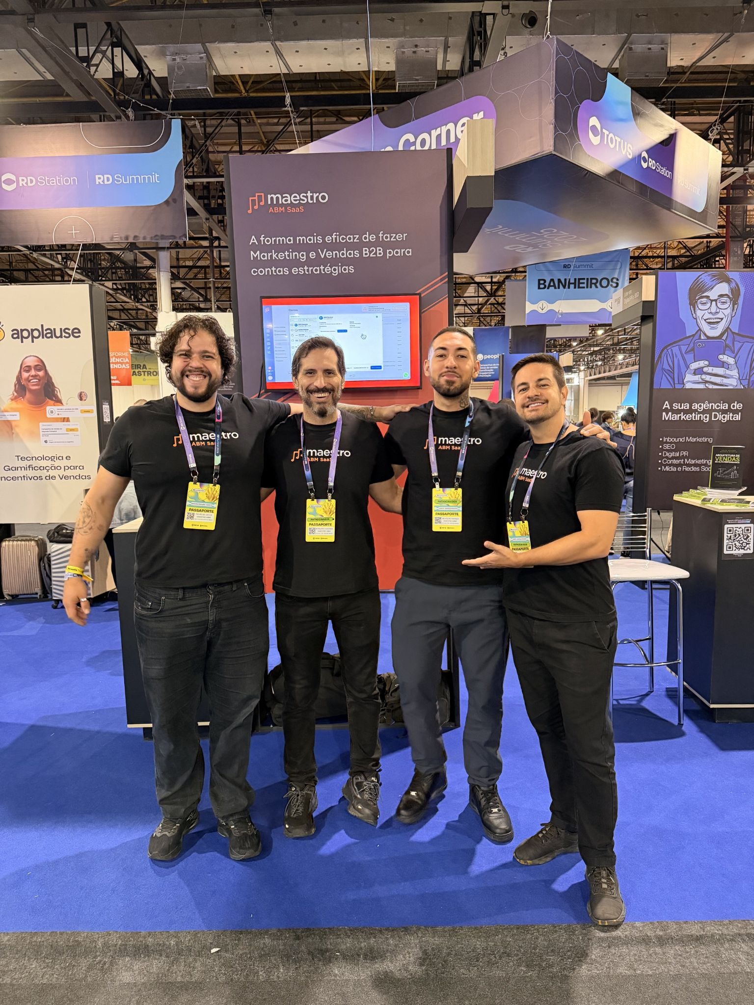 Four smiling men stand side by side at a tech expo, wearing matching "maestro" shirts. Background features colorful banners and a tech booth.