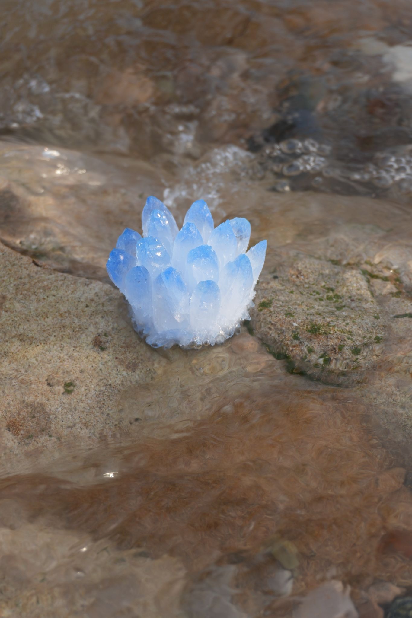 A cluster of blue crystals sits on a sandy, rocky surface by flowing water. The translucent crystals and gentle waves create a serene, natural scene.