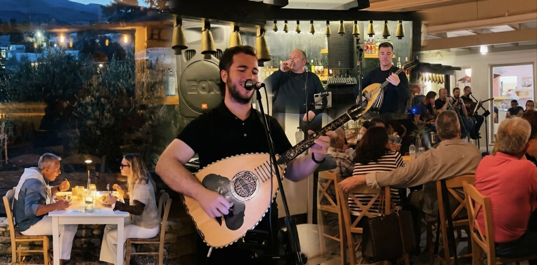 A man plays guitar in Souvlaki Dryos, a lively restaurant on Paros Island, with patrons enjoying their meals around him.
