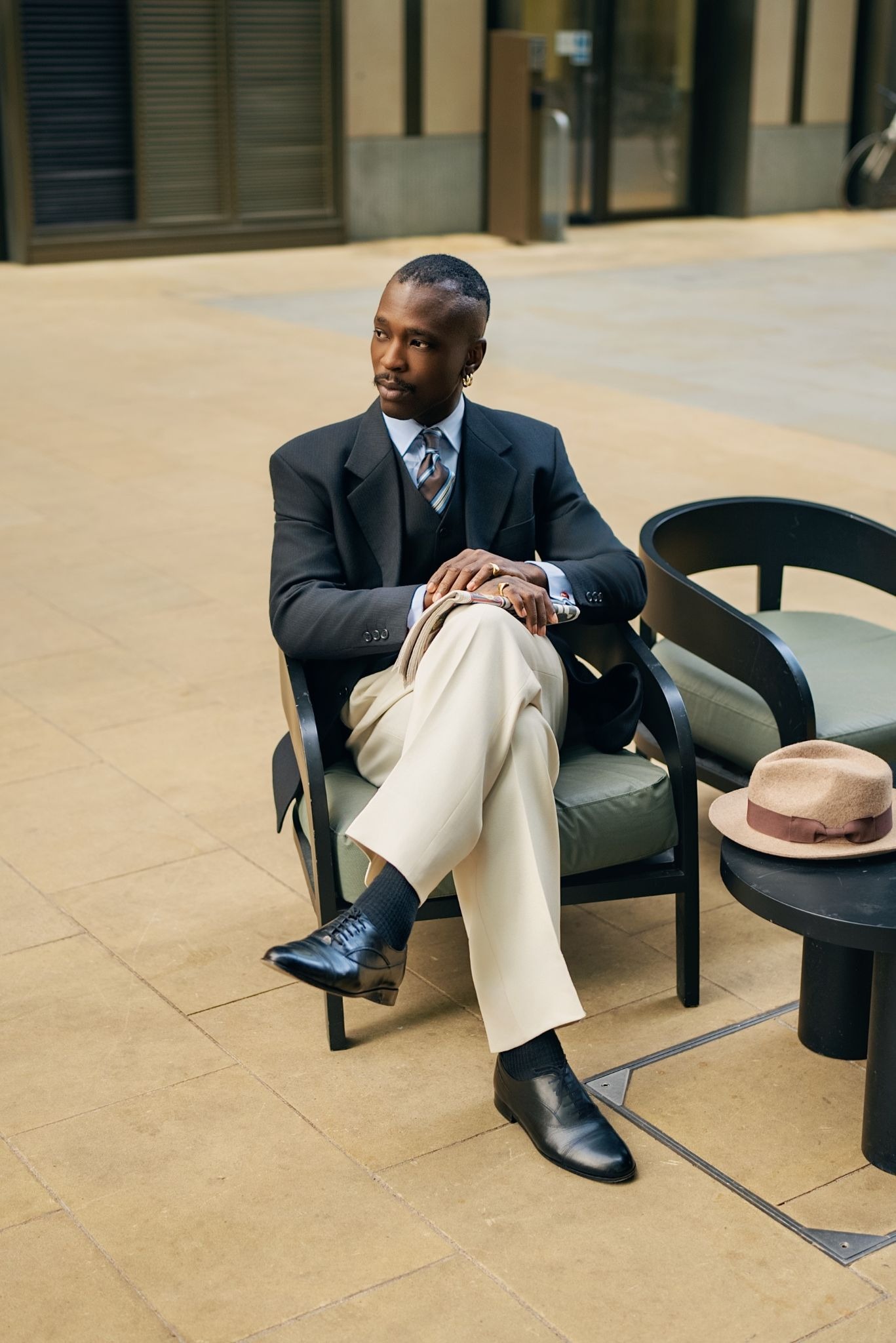 A man in a black blazer and light trousers sits outdoors, legs crossed, exuding confidence. A hat rests on a nearby chair. The mood is elegant and relaxed.