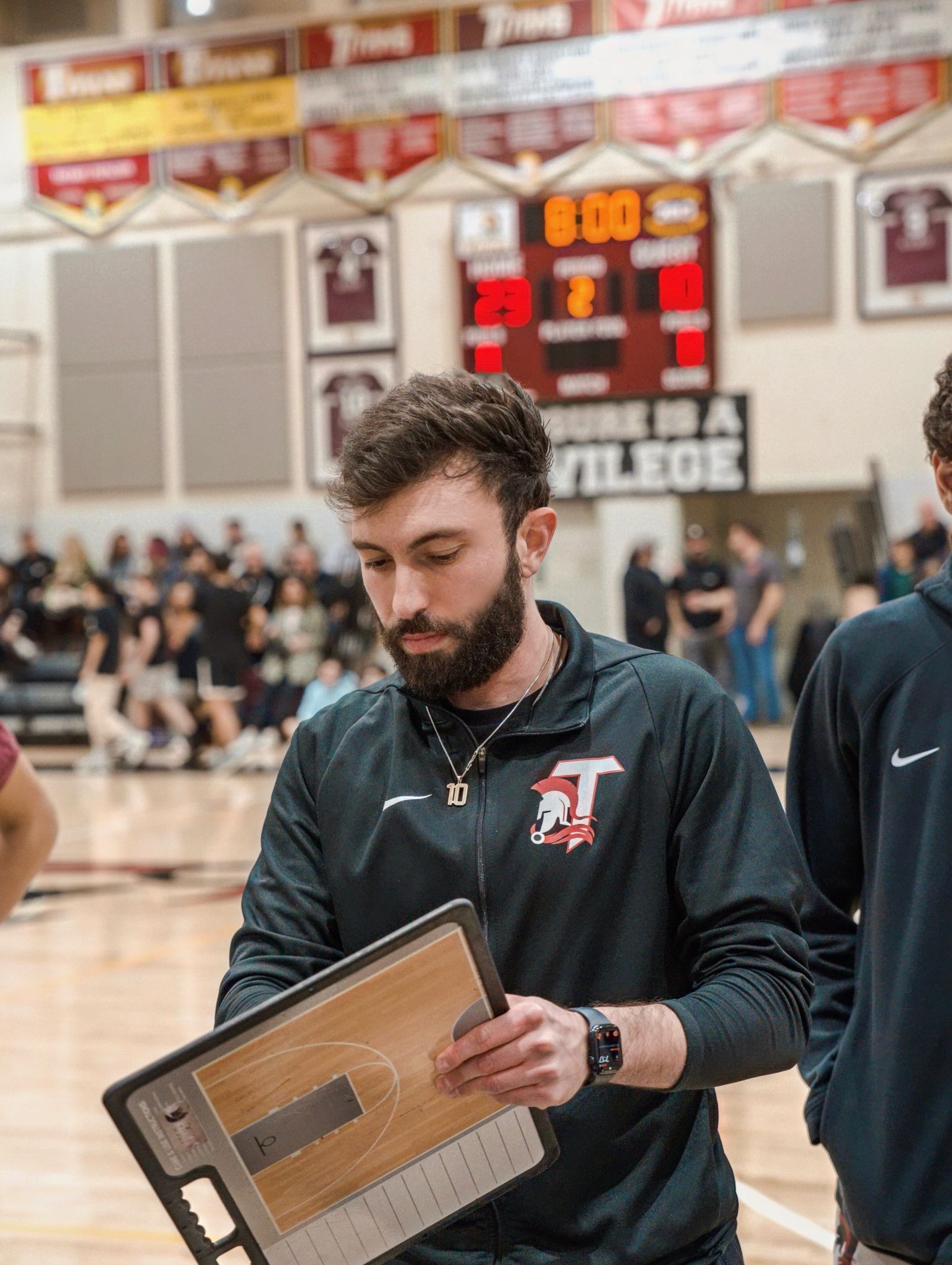 Coach standing on sideline addressing players during game with spectators in background.