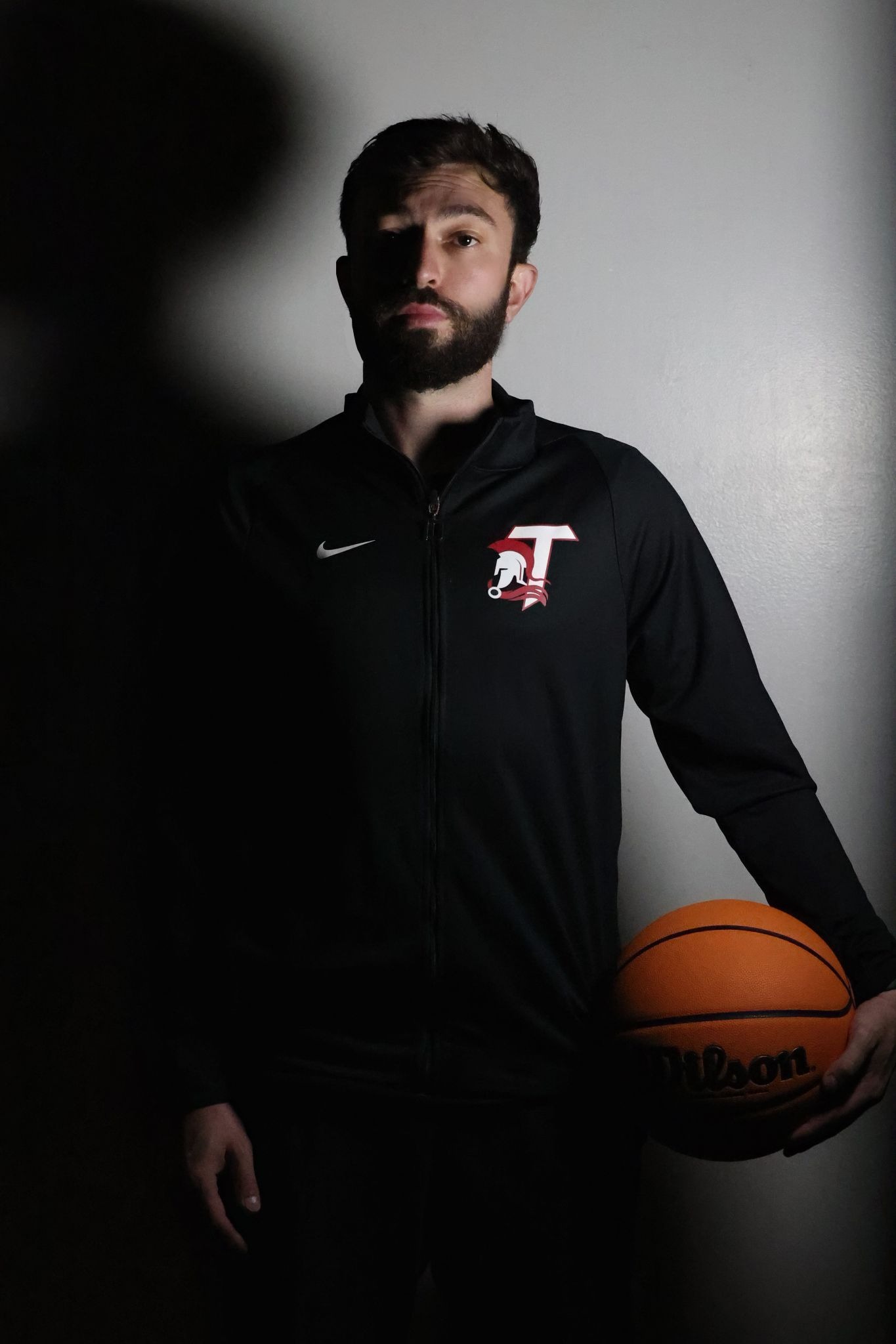 Coach holding basketball in dramatic low lighting against plain wall.