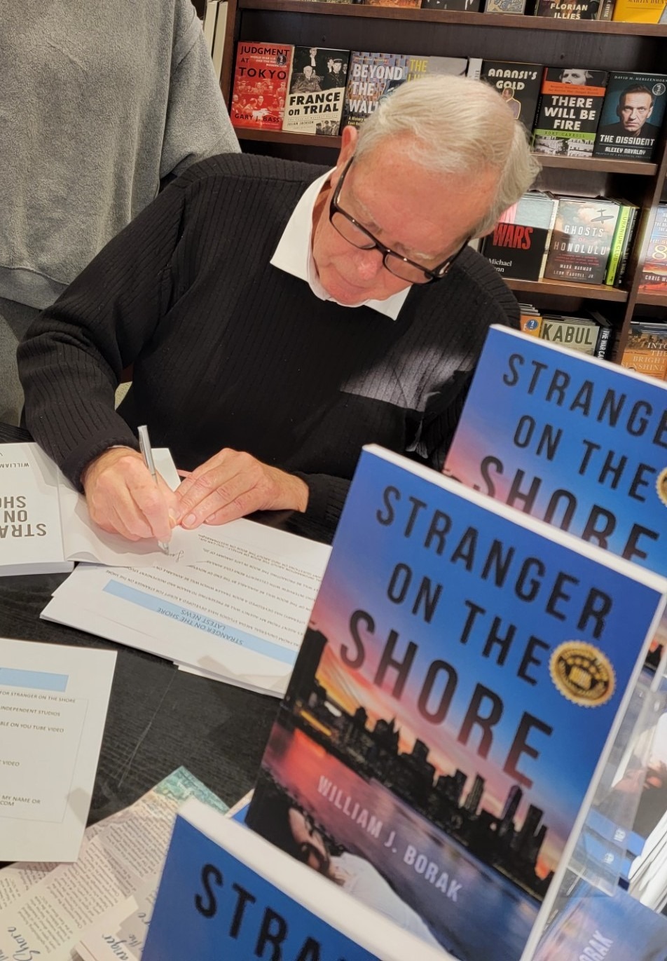A man with gray hair signs books titled "Stranger on the Shore" at a book signing event. Shelves in the background display various books.