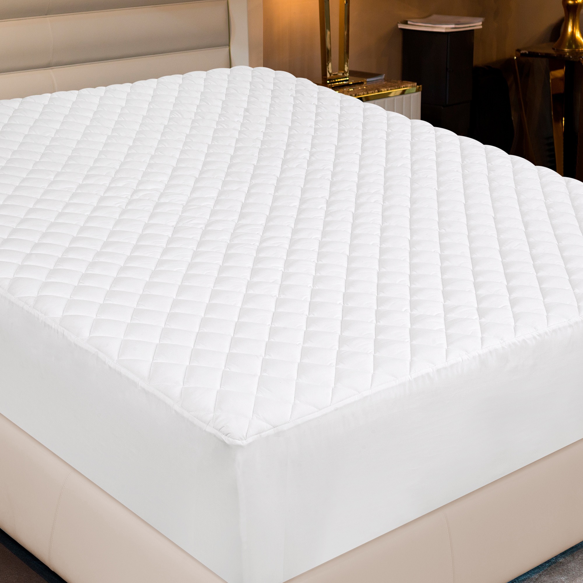 A white quilted mattress pad covers a bed in a cozy bedroom setting, evoking a sense of comfort and cleanliness. A bedside lamp and table are in the background.
