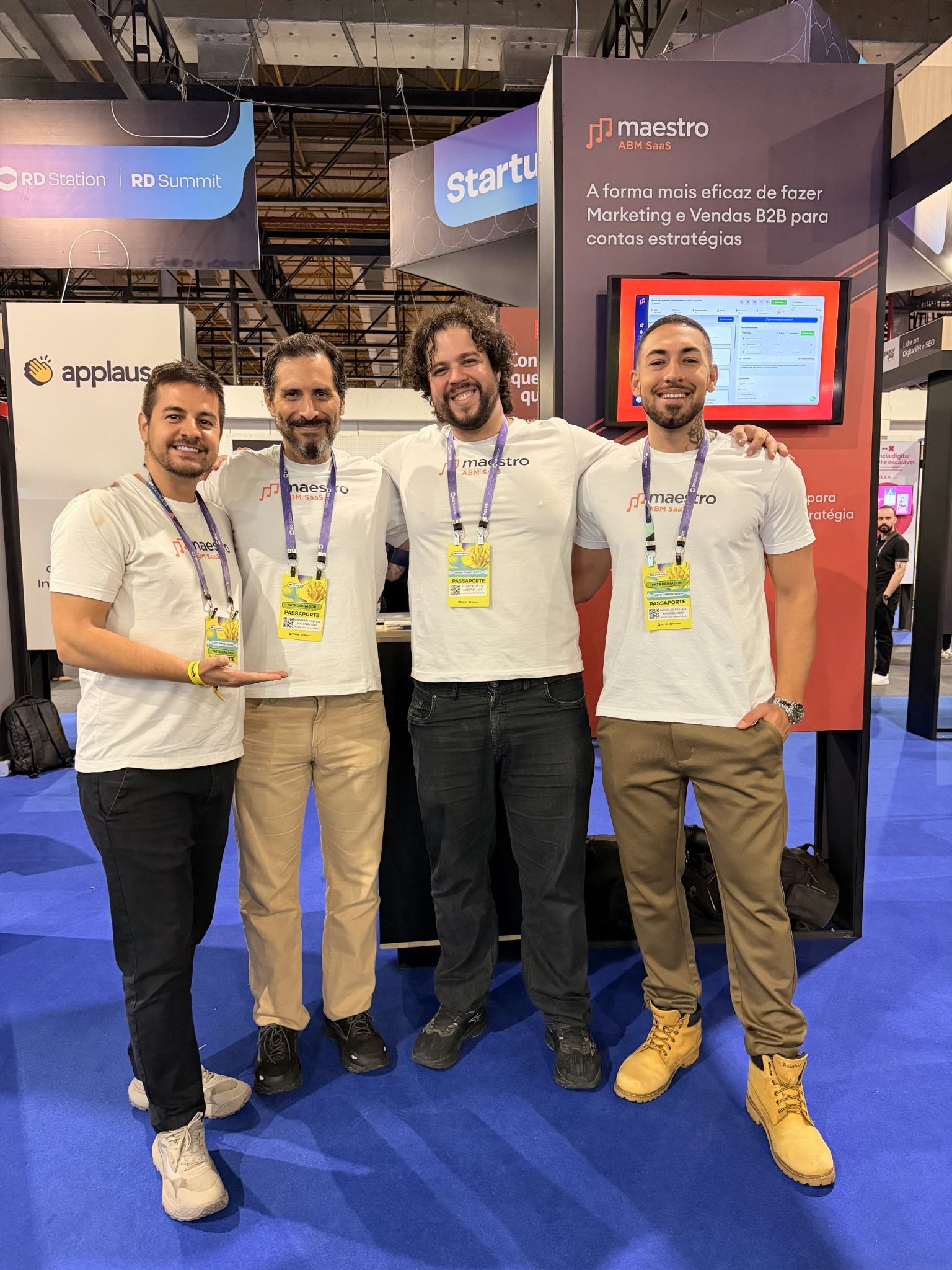 Four men wearing matching logo t-shirts stand smiling at an event booth with a blue carpet. They convey a friendly and professional atmosphere.