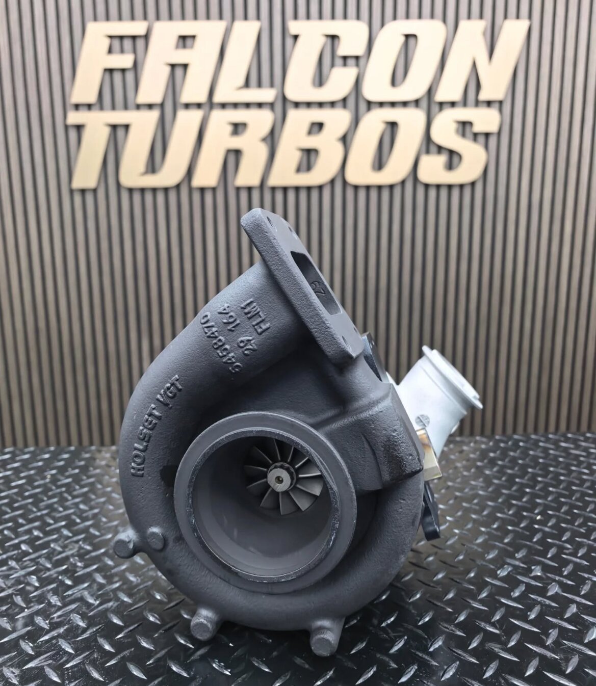 Black performance turbocharger with visible turbine blades positioned before Falcon Turbos workshop branding.