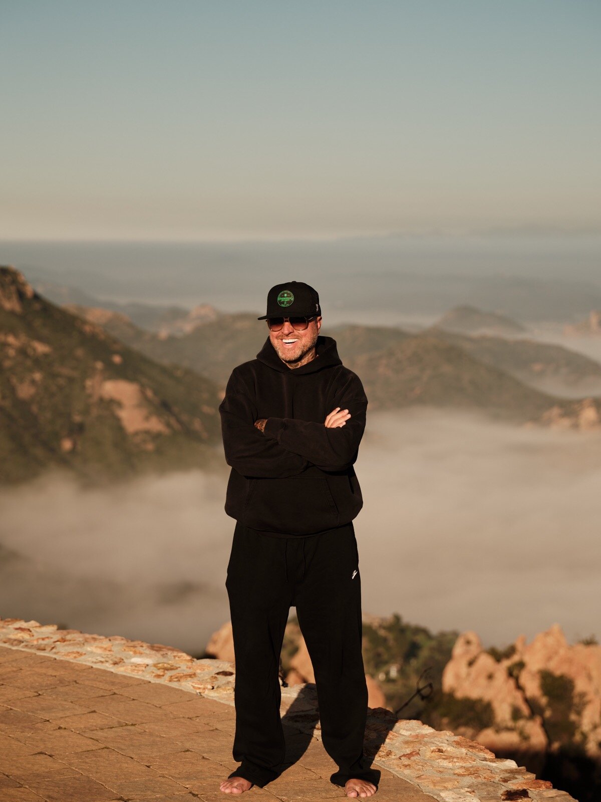 Brandon Johns, wearing a black sweatshirt and hat, stands on a mountain, representing his digital brand and entrepreneurial spirit.