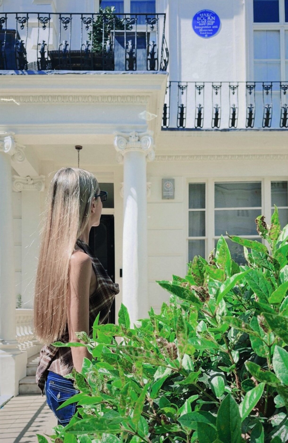 Long-haired woman standing beside greenery outside elegant white townhouse with balconies in bright sunlight.
