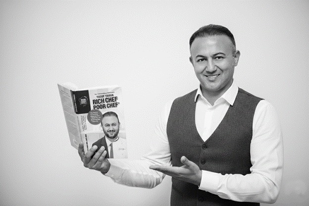 Yusuf Yaran smiling while presenting his book Rich Chef Poor Chef against a plain studio background.