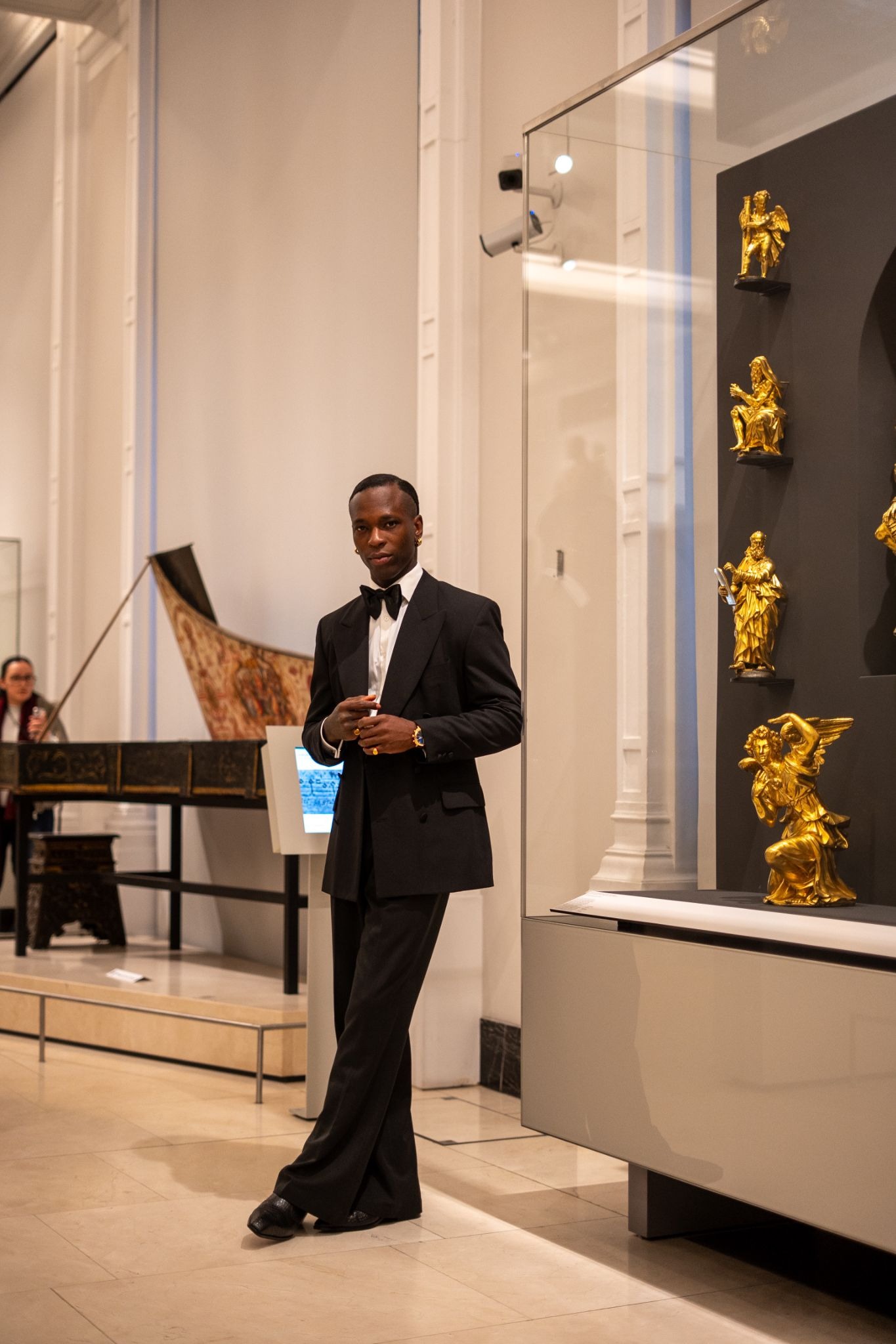 A well-dressed man in a black tuxedo leans against a museum display case with golden sculptures. The atmosphere is elegant and sophisticated.