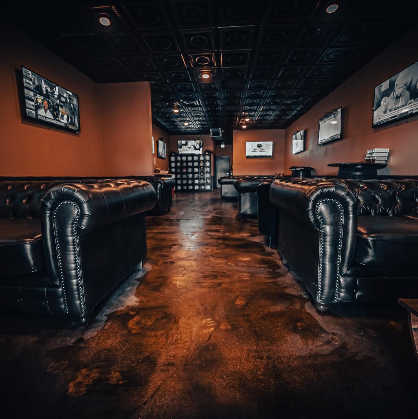 Dimly lit lounge with black leather couches lining both sides, walls adorned with televisions, and a dark, polished floor, creating a cozy, sophisticated ambiance.