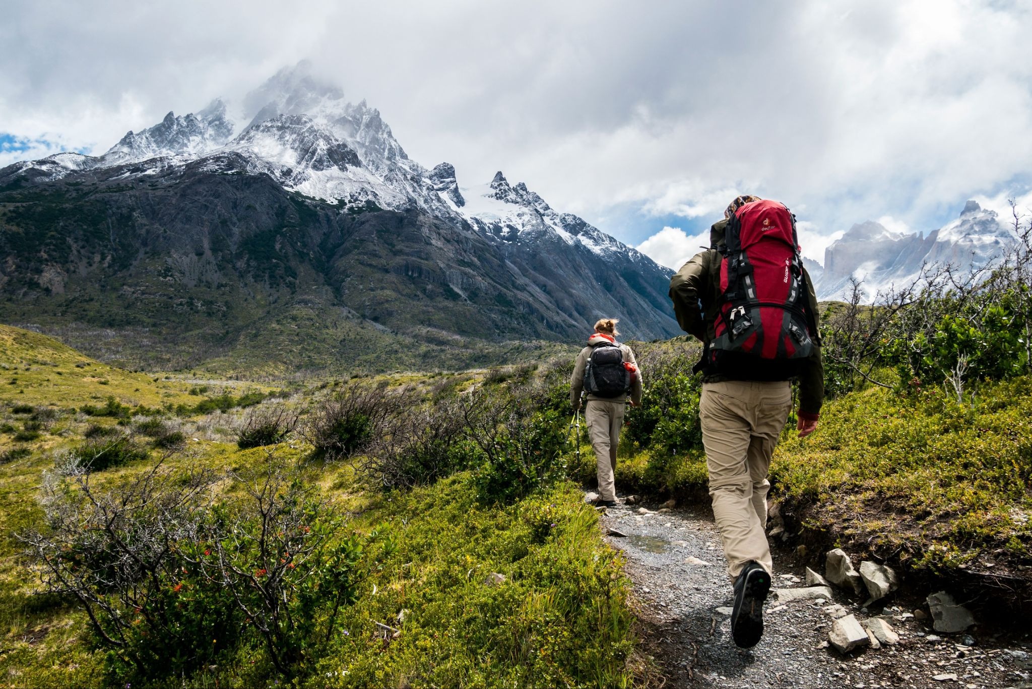 Hikers trekking along mountain trail beneath snowcapped peaks.