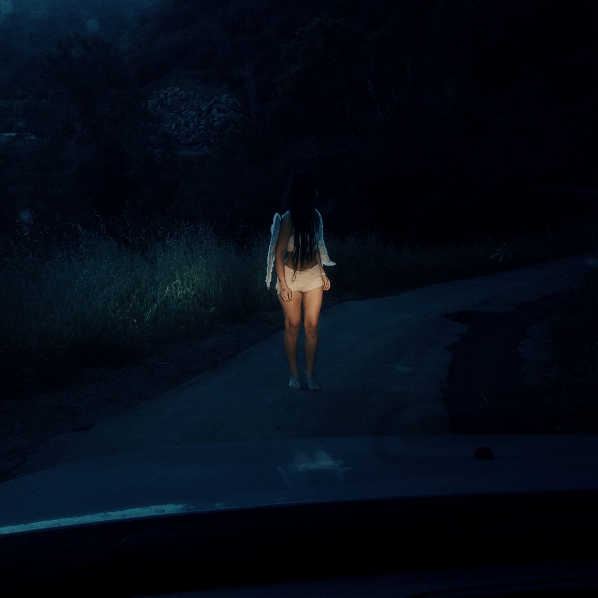 Person with angel wings standing barefoot on dark rural road illuminated by headlights at night.
