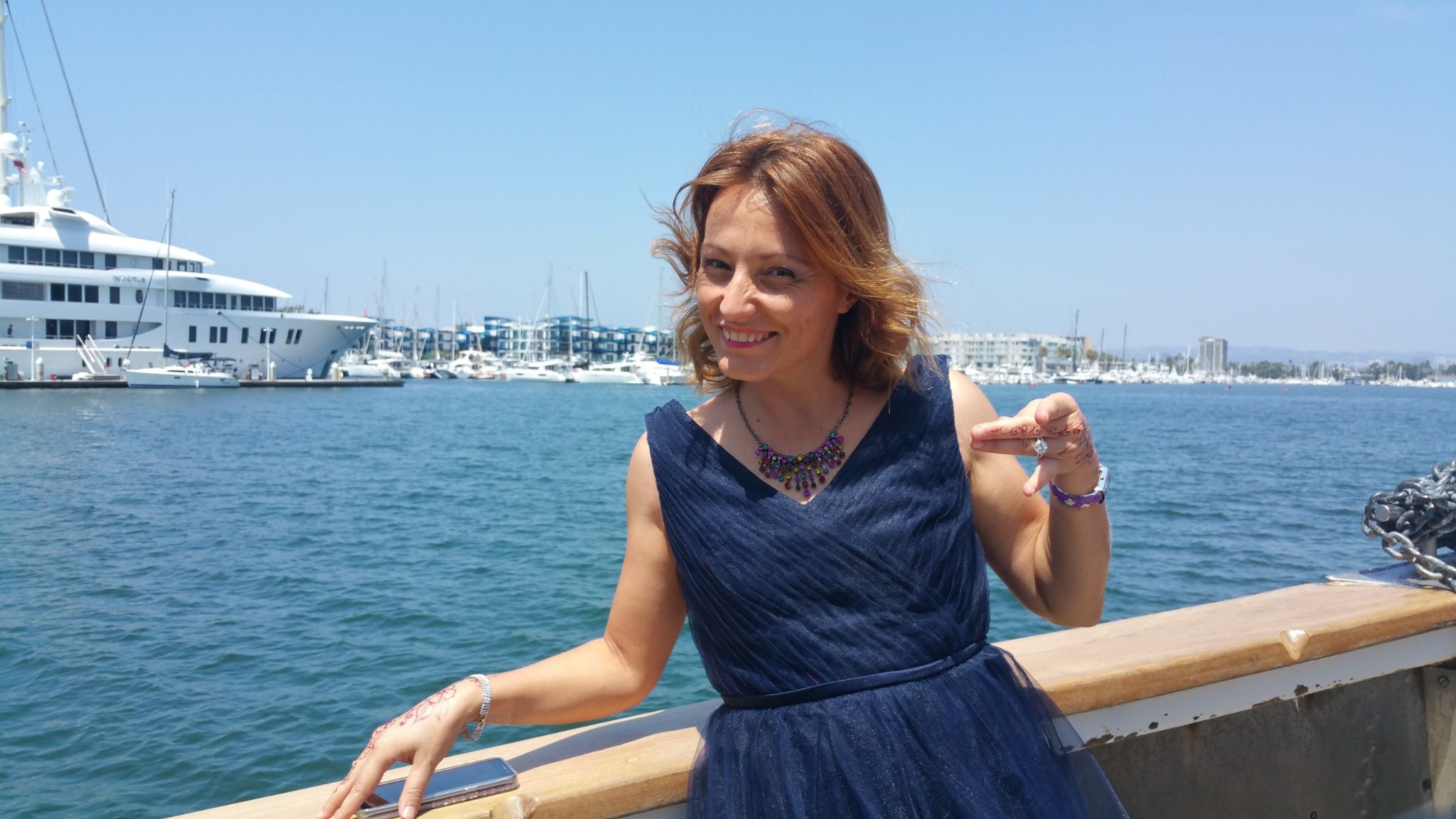 Woman in navy dress smiling by marina, pointing at engagement ring with yachts and waterfront skyline behind her.
