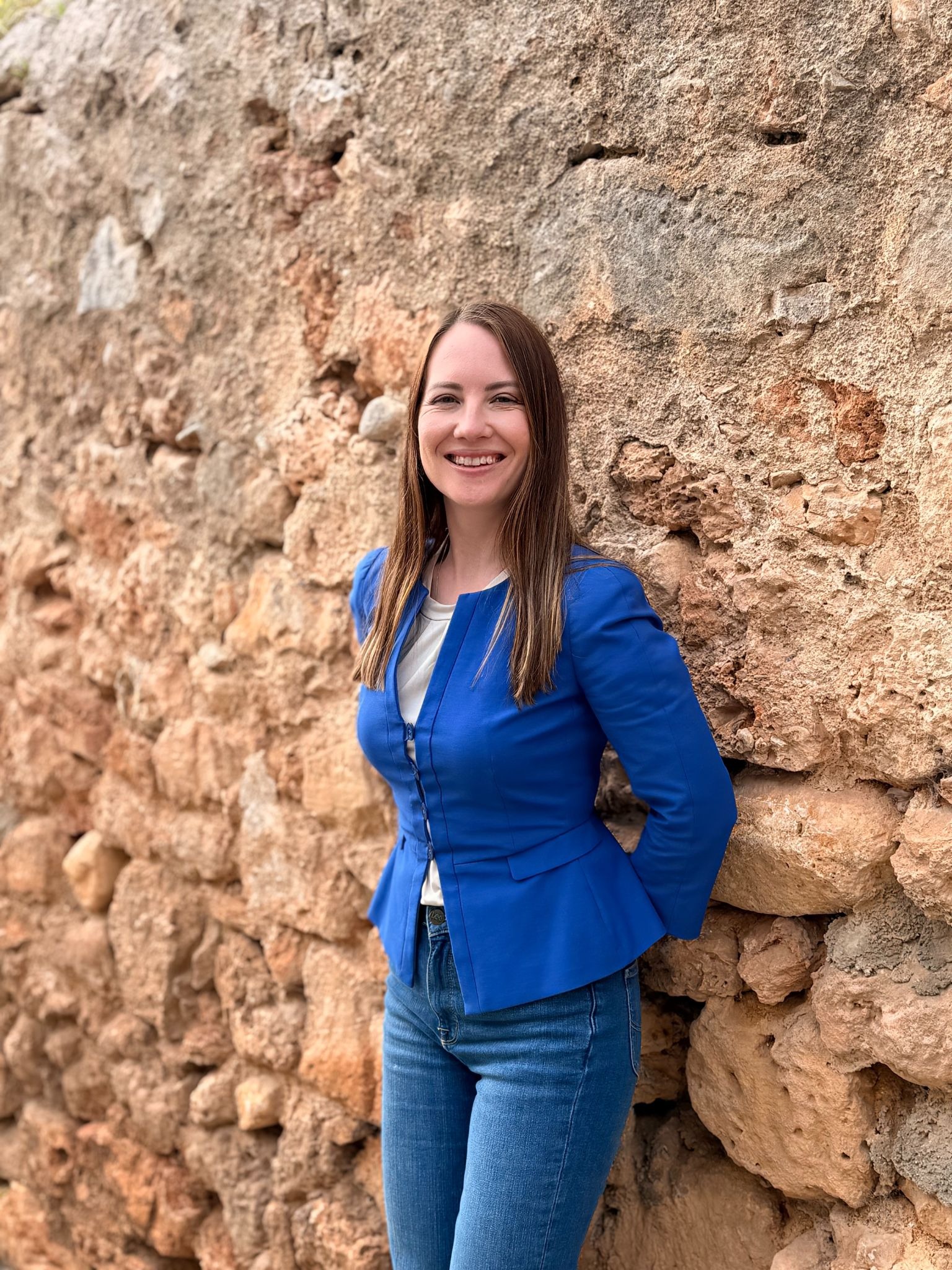 Woman in blue blazer smiling beside rustic stone wall outdoors.`