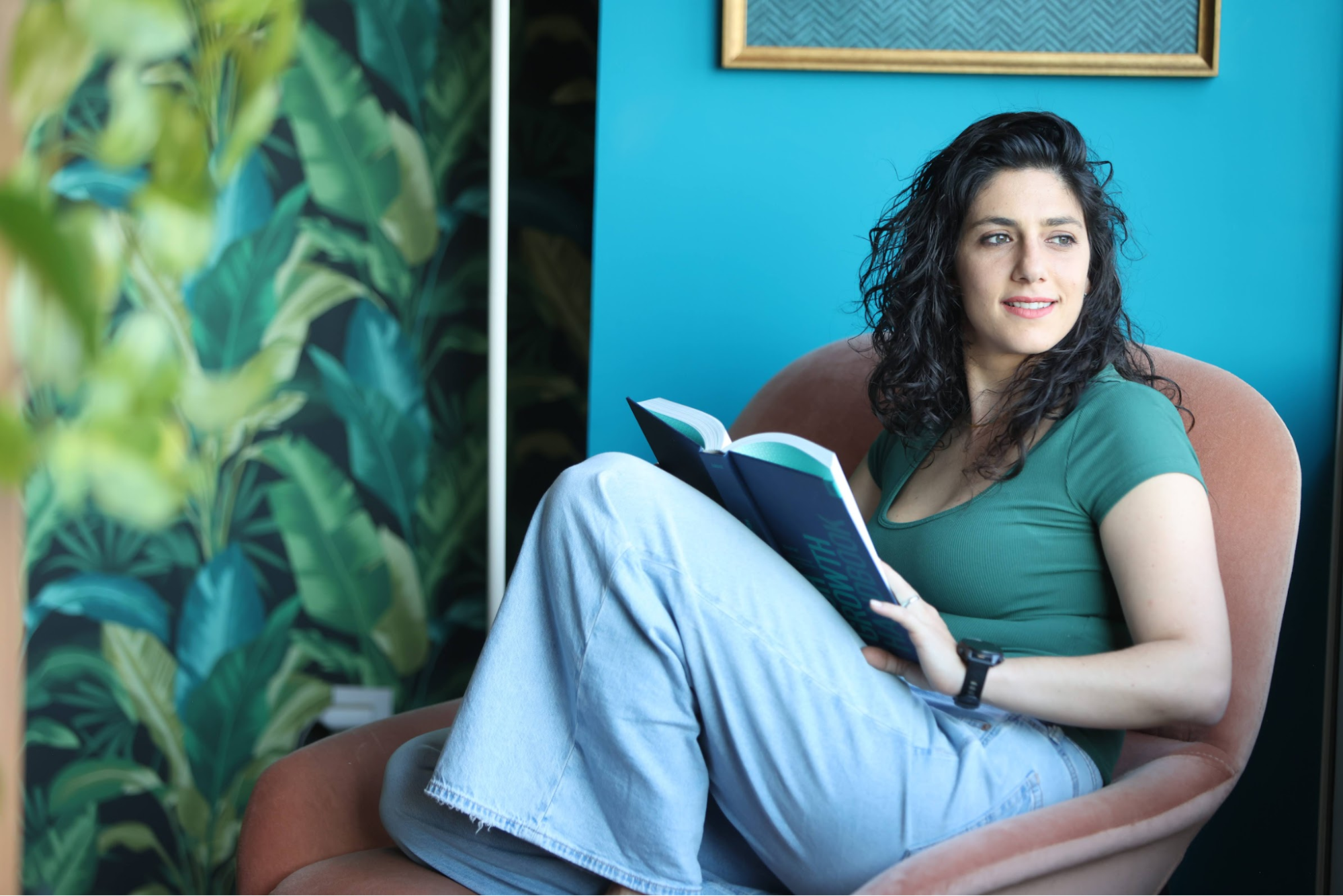 Woman relaxing in a chair reading a book in a modern, plant-filled room.