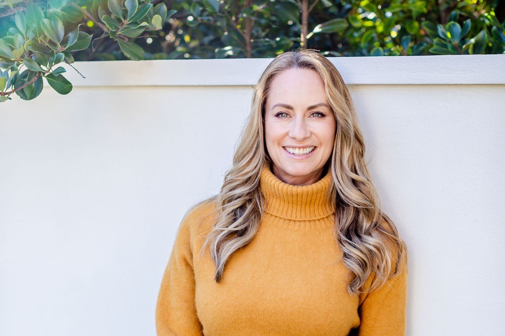 Professional woman in mustard turtleneck smiling outdoors against a white wall with greenery.