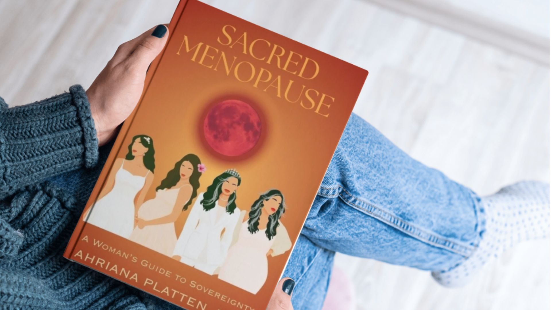 Woman holding “Sacred Menopause” book while sitting casually in jeans and a knitted sweater.