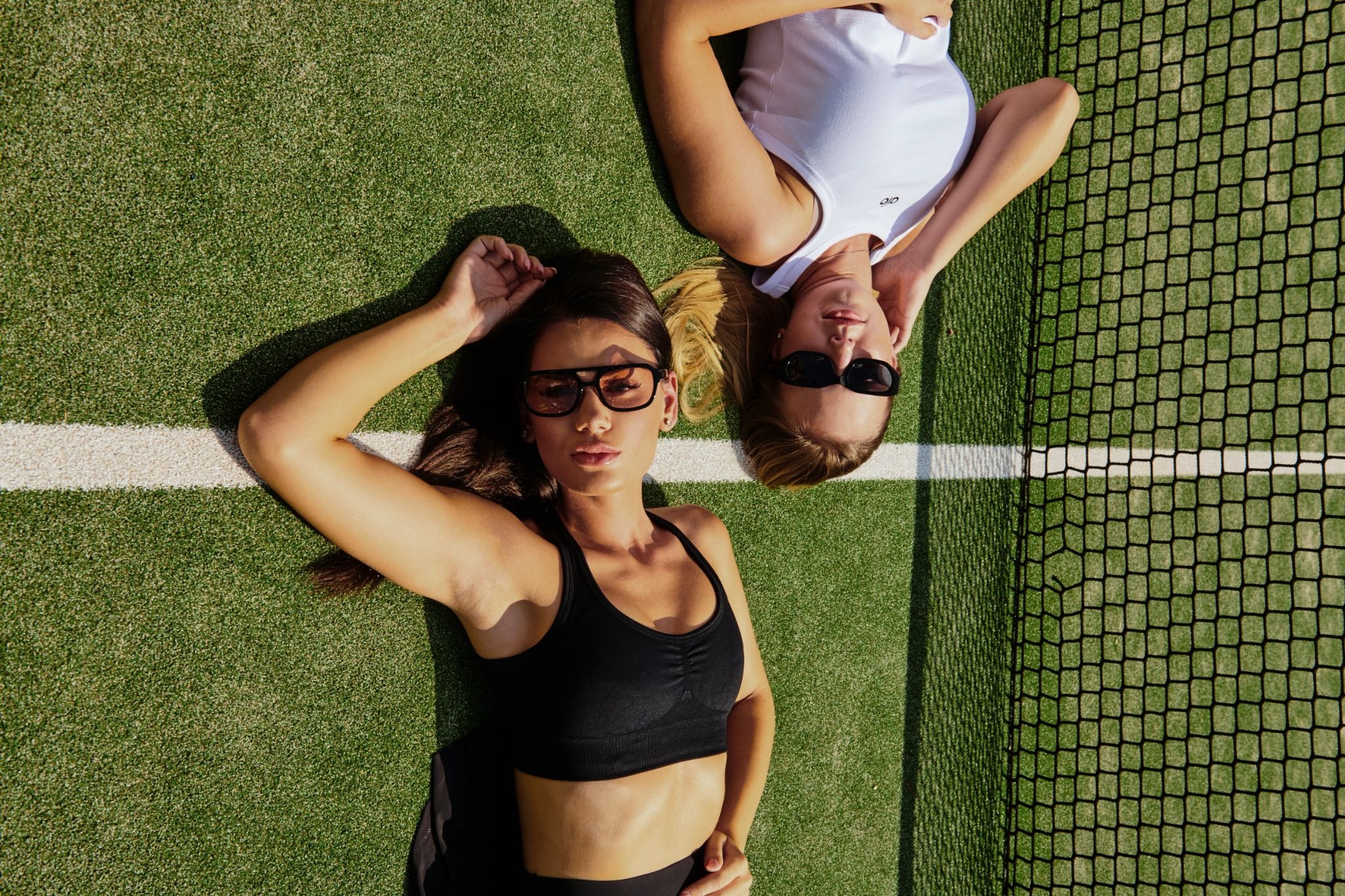Two women in sports attire lie on a tennis court near the net, wearing sunglasses. Their relaxed pose and attire suggest a sporty, casual vibe.