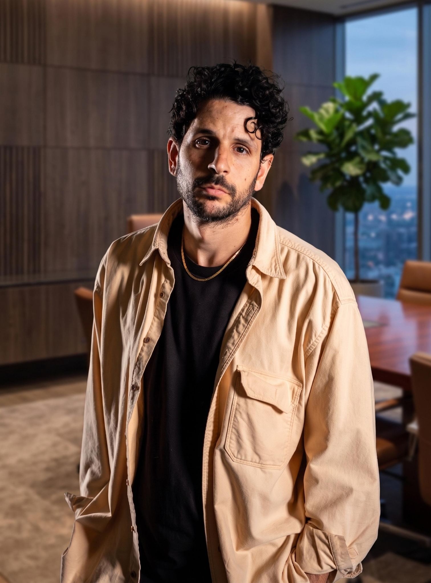Man in a beige jacket, standing in an office with a plant in the background.