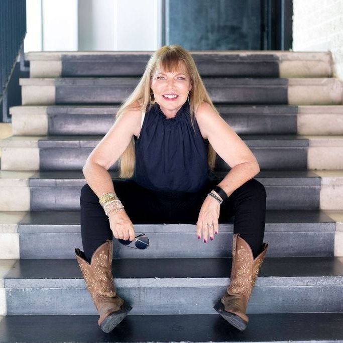 Karel Glazer seated on indoor steps, smiling, wearing a black sleeveless top and cowboy boots.